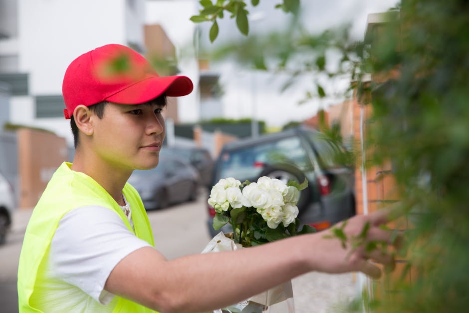 A young male flower delivery courier wearing a red cap and high-visibility yellow vest is seen outdoors on a residential street, carefully handing over a bouquet of fresh white flowers. The bouquet fe