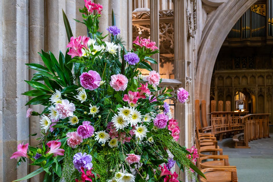 A vibrant bouquet arrangement of fresh flowers placed inside a church, featuring pink, purple, and white blooms including lilies, carnations, and daisies, accented with lush green foliage. The flowers