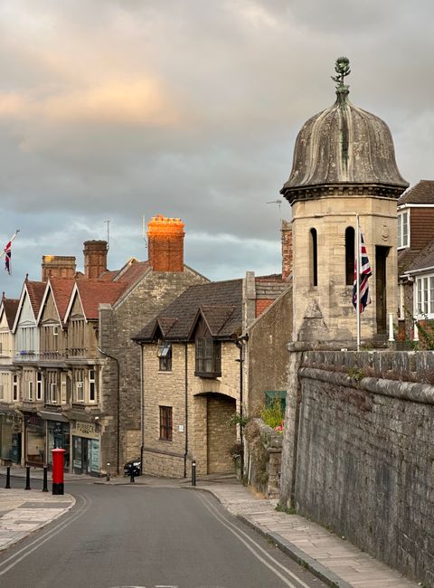 The image depicts a quaint street scene in Chalford Saint Giles with historic stone buildings featuring pitched roofs and chimneys, set against a cloudy sky during the late afternoon or early evening.