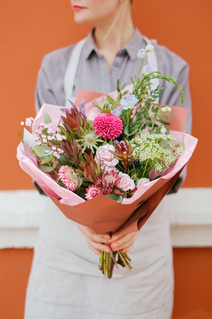 A person holding a vibrant bouquet of fresh flowers arranged with various pink, white, and purple blooms, including gerbera daisies, roses, and carnations, complemented by green foliage. The bouquet i