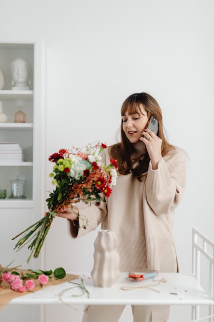 A woman with long brown hair and a beige sweater is arranging a vibrant bouquet of fresh flowers, including red, white, pink, and green blooms, on a white table inside a well-lit room. She is holding 