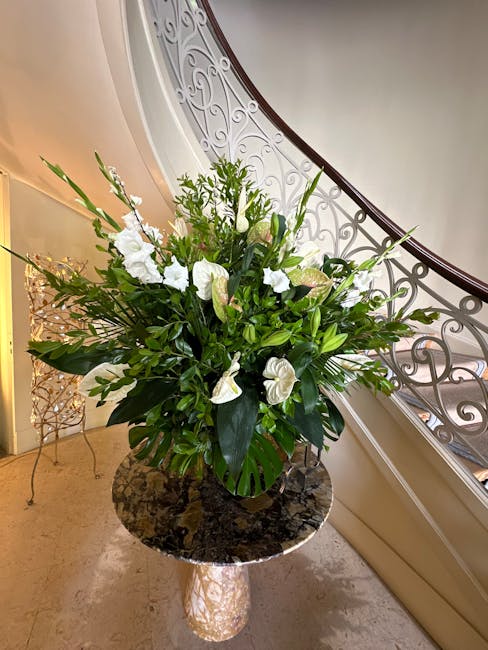 A large floral arrangement placed on a polished, dark marble-topped side table near a curved staircase with ornate iron balustrades. The bouquet features white lilies, anthuriums, and blooms with soft