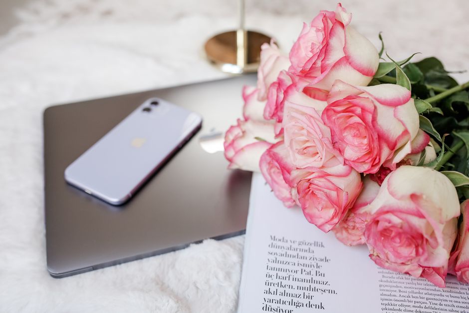 A floral arrangement of fresh pink and white roses with delicate petals and green leaves, resting on a white surface alongside a silver laptop and an iPhone. The roses are partially wrapped in white p