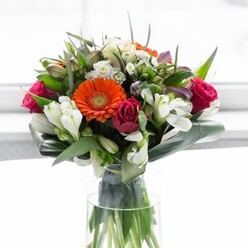 This exquisite bouquet, arranged with the artistry honed by Chalfont St Giles florists, stands tall and graceful in a clear vase, reflecting the gentle balance of colour and delight found throughout the village. The creamy peach and bright orange gerberas form a radiant ring of light, recalling the sun-warmed afternoons on Narcot Lane. Deep fuchsia roses rise at their side, their rich hues layered with romance and tradition that speaks to the area's centuries-old heritage. Between bold blooms, lacy clusters of white alstroemeria billow like soft clouds, touched by glimmers of pink and whispers of airy white filler flowers-a nod to the abundant florals that spill from local hedgerows. Broad, paddle-shaped green leaves arch securely around the arrangement's base, creating a lush, protective collar much like the shelter of ancient oaks on the Green, while smaller pointed leaves and trailing tendrils add vibrancy and dimension. Light pours in from behind, highlighting the joyful spectrum of color and the bouquet's sculptural beauty, while the clear vase reveals verdant green stems that emphasize the arrangement's freshness and integrity. Imbued with a subtle sense of place and celebration, this bouquet feels ready for sharing with cherished friends, enlivening family gatherings, or gifting heartfelt thanks throughout Chalfont St Giles and its countryside surrounds.